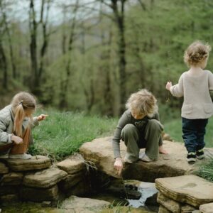 Three children playing by a stone-lined stream in a lush green forest during springtime.