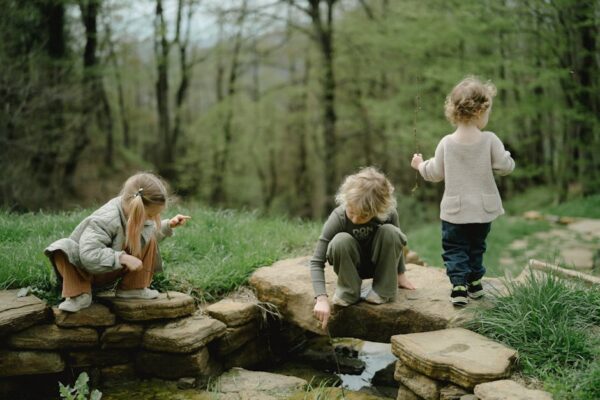 Three children playing by a stone-lined stream in a lush green forest during springtime.
